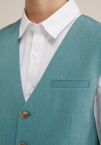 Tyrkysová vesta s hladkým povrchem, pod kterou se nachází bílá košile s knoflíky. Na hrudi je jedna kapsa a vesta má hnědé knoflíky.