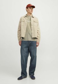 Beige denimjakke over en grøn t-shirt med korte ærmer, parret med løse blå jeans og marineblå sneakers. Modellen bærer en rød kasket.