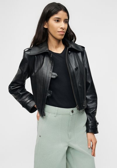 Νεαρή γυναίκα στέκεται με το χέρι στην τσέπη φορώντας μαύρο δερμάτινο μπουφάν πάνω από μαύρο τοπ και ανοιχτοπράσινο παντελόνι ψηλόμεσο, σε λευκό φόντο.