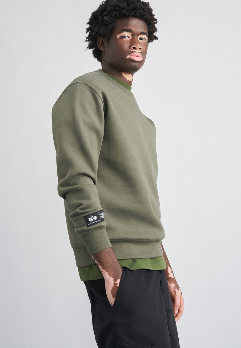 Φούτερ σε ελαιί πράσινο με λεία υφή, στρογγυλή λαιμόκοψη και μανίκια με μανσέτα. Διαθέτει μαύρη ετικέτα στο αριστερό μανίκι και από κάτω φορεμένο ένα στρωματοποιημένο πράσινο πουκάμισο.