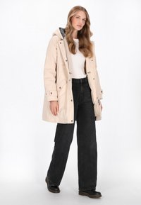 Lys beige vandtæt parka med hætte, trykknapper og sidelommer, parret med høj talje sorte jeans og sorte støvler.
