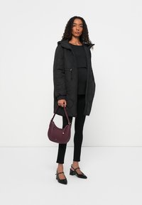 Abrigo acolchado negro con capucha, parte superior negra y leggings, combinado con un bolso de hombro burdeos y zapatos de tacón negro con punta.