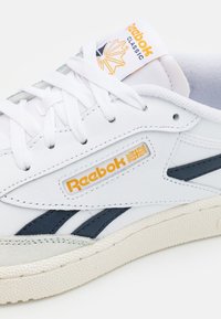 Sneaker di pelle bianca con dettagli blu navy, suola testurizzata e logo Reebok arancione. Presenta un design con lacci e una linguetta imbottita.