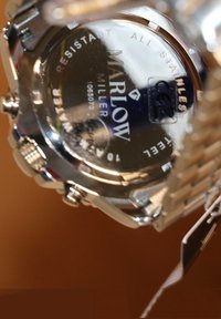 Stainless steel watch with a reflective silver face, marked "Marlowe Miller," featuring water resistance and a brushed metal band.