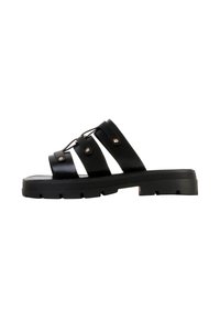 Sandale à semelle épaisse noire avec trois larges brides ornées de clous métalliques et un design à lacets sur fond blanc.