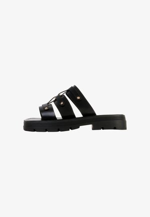 Sandalia con suela negra gruesa y tres tiras anchas con detalles de tachuelas metálicas y diseño de cordones sobre un fondo blanco.