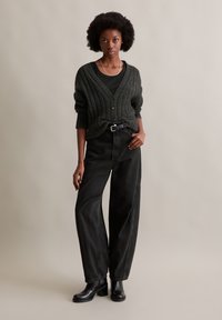 Donkergroen geribd cardigan met knopen, gecombineerd met een zwarte top en losse zwarte broek. Zwarte enkellaarsjes maken de outfit compleet.