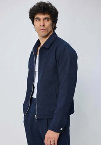 Veste zippée bleu marine en tissu doux, dotée d'un col, de poches latérales et d'accents en bouton sur les manches. Associée à une chemise blanche.