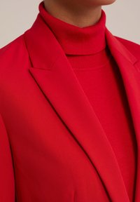 Röd kavaj med skarpa slag, kombinerad med en röd polotröja. Slät, strukturerad tyg med en figurnära design förstärker den livliga färgen.