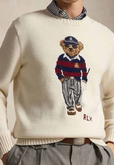 Homme portant un pull en maille crème avec un ours brodé sur une chemise de rugby rayée, casquette, lunettes de soleil et tenue de golf tenant un club.