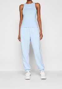 Kvinna iklädd en ljusblå Adidas-topp och joggers, matchade med vita sneakers, stående framför en ljus och enfärgad bakgrund.