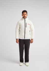 Giacca a vento bianca con cappuccio e texture trapuntata orizzontale, con zip frontale e tasche laterali, abbinata a pantaloni neri e sneaker bianche.