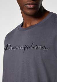 Graues Baumwoll-T-Shirt mit Rundhalsausschnitt, das mit einem gestickten "Champion"-Logo in einem dunkleren Farbton versehen ist. Glatte Textur mit lockerer Passform.