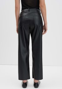 Vrouw in zwarte hoge taille nepleer wijde broek en zwarte instap schoenen, gezien vanaf de achterkant tegen een effen achtergrond.