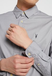 Mężczyzna zapięty guzik mankietu na jasnoszarej koszuli z małym czarnym logo czaszki zwierzęcia na piersi.