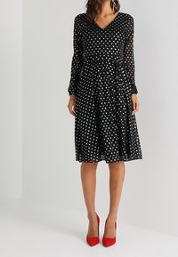 Schwarzes Pünktchenkleid mit V-Ausschnitt, langen transparenten Ärmeln und einem Bindegürtel, das einen fließenden Rock und rote High Heels hat.