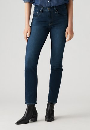 Femme portant un jean slim bleu foncé, des bottines noires et une chemise en denim rentrée dans le pantalon, sur un fond clair uni.