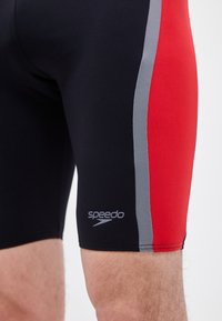 Άνω μέρος των ποδιών άνδρα που φοράει μαύρα μαγιό με κόκκινες και γκρι πλαϊνές πινακίδες και λευκό λογότυπο Speedo στο αριστερό μηρό.