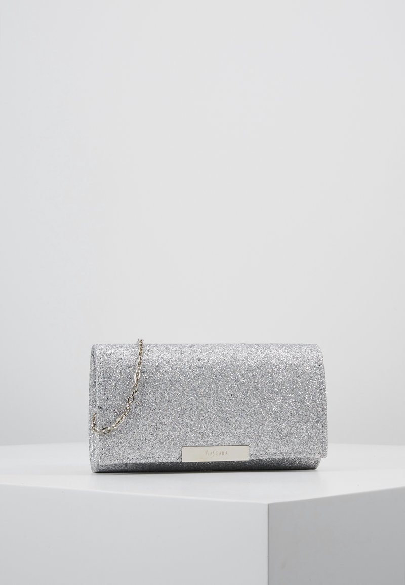 Pochette pailletée argentée avec une petite bandoulière chaîne, posée sur une surface blanche devant un fond gris clair uni.