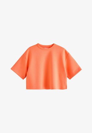 Barn med krøllet hår iført en oversized orange skjorte og lyse denimbukser, stående mod en ensfarvet beige baggrund.