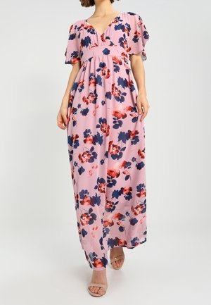 Vrouw draagt een roze bloemen maxi-jurk met korte mouwen en een diepe V-hals, gecombineerd met beige sandalen met open teen.