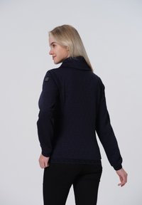 Veste matelassée marine avec un col haut, ornée d'un motif triangulaire et d'une bande décorative à l'ourlet, fabriquée en tissu léger.