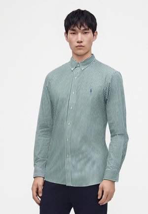 Jeune homme portant une chemise à rayures verticales vertes et blanches avec des manches longues et un pantalon sombre, debout devant un fond blanc uni.