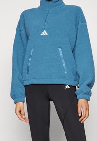 Fleece-pullover i blått med hög krage, dragkedja och två sidofickor. Har en vit Adidas-logotyp och satinbanddetaljer.