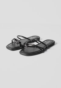 Schwarze flache Sandalen mit quadratischen Zehen, dünnen Riemen und dekorativem Kettendetail vorne, dargestellt vor grauem Hintergrund.