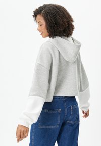 Gekürzter grauer Hoodie mit weißen Ärmeln, aus weichem Stoff gefertigt. Verfügt über eine Kapuze und einen lockeren Schnitt, kombiniert mit blauen Jeans.