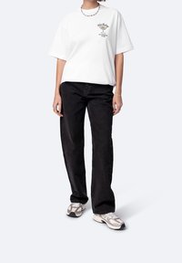 Biała, oversize'owa koszulka z nadrukiem graficznym, w połączeniu z czarnymi jeansami typu straight-leg. Obuwie to jasne buty sportowe.