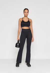 Svart idretts crop top og vide tights laget av teksturert stoff, sammen med svarte idrettssko og holder en svart kettlebell.