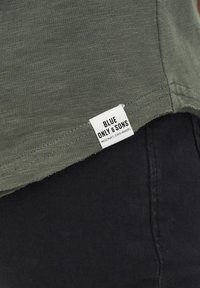 Olivgrünes Hemd mit weicher Textur, mit einem schwarzen Saum und einem weißen Etikett, auf dem "BLUE ONLY & SONS" sowie Pflegehinweise stehen.