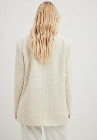 Teksturoidettu vaalean kerma takki, jossa on räätälöity muotoilu, hieman ylisuurena ja huomaamattomilla saumoilla. Kevyt kangas, jossa on hienovaraisia kohotettuja kuvioita.