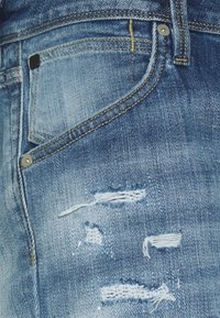 Blå denimjeans med en klassisk design, slitna detaljer med trasiga lappar och metallbeslag nära fickorna.