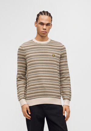 Ung mand med fletninger iført en beige mønstret sweater med lille gult logo på brystet og sorte bukser, stående foran en ensfarvet baggrund.