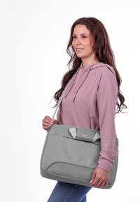 Frau in pinkem Hoodie, die eine graue Umhängetasche mit Griffen hält, vor einem weißen Hintergrund steht, nach vorne schaut und lächelt.