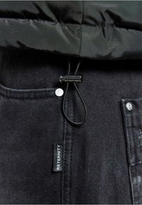 Sorte denimjeans med et kontrastfarvet vævet label, metalknapdetaljer og snor i taljen. Teksturen er glat og robust.