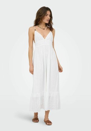 Femme portant une robe maxi blanche sans manches avec un décolleté en V profond et un ourlet à volants, accompagnée de sandales slides marron et d'un collier de perles.