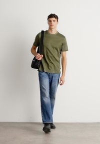 Moški hodi v olivno zeleni majici, modrih kavbojkah, črnih čevljih in nosi črno torbo za čez ramo, proti enostavni beli ozadju.