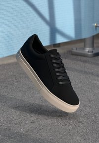Sneaker negru cu un superior din țesătură netedă, șireturi negre plate și o talpă din cauciuc bej deschis, având un design simplu și o formă rotunjită.