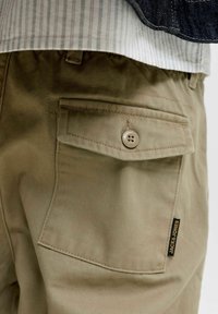 Pantalon kaki avec une poche à rabat boutonnée et une étiquette de la marque Jack & Jones sur le côté, porté avec une chemise rayée et une veste en jean.