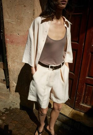 Femme portant un short ample couleur crème et une chemise ouverte assortie sur un débardeur taupe, debout à l'extérieur près d'un mur texturé et d'une porte en bois.