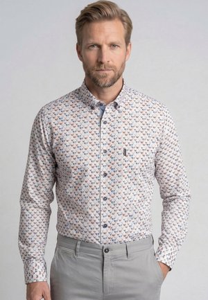 Man met lichtbruin haar en baard draagt een patroon lange mouwen overhemd met knopen en lichtgrijze broek met één hand in de zak.