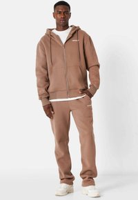 Felpa con zip marrone e logo bianco, abbinata a pantaloni della tuta marroni coordinati. Entrambi sono realizzati in un morbido tessuto lavorato. Sneakers bianche completano il look.