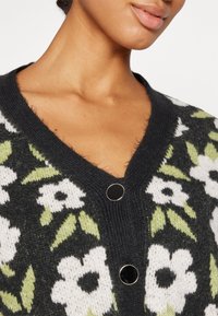 Stickad cardigan med mörk bakgrund, prydd med vita blommor och gröna blad. V-ringad design med svarta knappar som utmärkande detaljer.