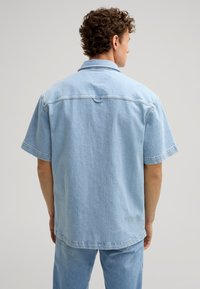 Camicia in denim azzurro chiaro a maniche corte con vestibilità rilassata, colletto con bottoni e una singola cucitura nella parte posteriore. Texture liscia, design minimalista.