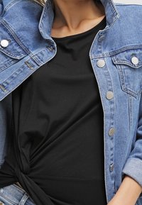 Žena v černém uzlu topu pod rozepnutou světle modrou džínovou bundou se stříbrnými knoflíky.