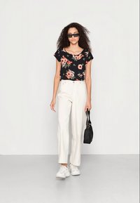 Camisa preta floral de mangas curtas, calças brancas e sapatilhas brancas; a segurar uma pequena mala preta. Texturas de tecido suaves.