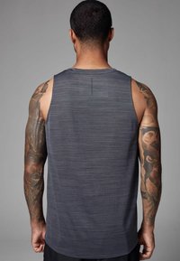 Homme aux cheveux courts avec des tatouages sur les bras entiers, portant un débardeur de sport gris foncé, tourné de dos à la caméra sur un fond gris.